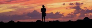 Silhouette of a person standing on rocks at sunset, with a vibrant orange-pink sky and scattered clouds above.